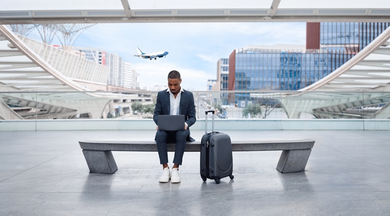 Young Black Businessman Using Laptop While Sitting On Bench At Modern Airport Terminal